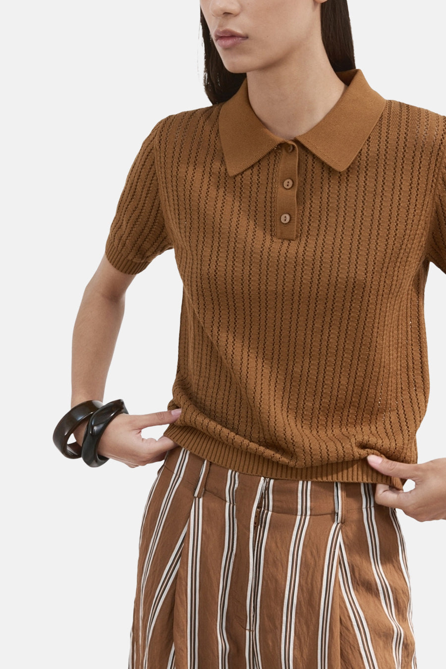 Bruine polo van MANGO, gedragen met een bruin-witte gestreepte broek en zwarte armbanden.