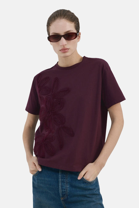 T-shirt à manches courtes bordeaux de MANGO, orné d'un motif floral ton sur ton, porté avec un jean et des lunettes de soleil.
