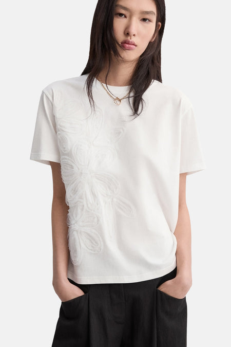 T-shirt blanc à manches courtes de MANGO, orné de broderies florales ton sur ton, porté avec un pantalon noir.

