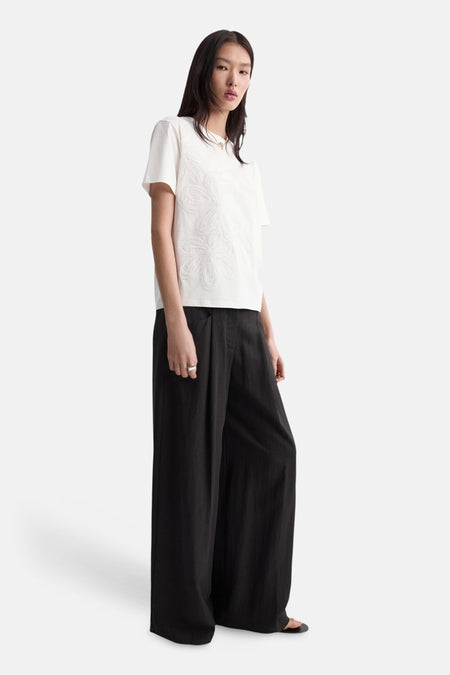T-shirt blanc à manches courtes de MANGO, avec motif floral ton sur ton, porté avec un pantalon noir ample.