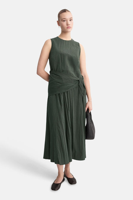 Robe kaki de MANGO avec une texture plissée, une ceinture nouée à la taille et des chaussures plates noires.

