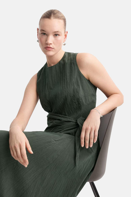Robe kaki de MANGO, portée par un mannequin assis, avec une ceinture nouée à la taille et des boucles d'oreilles discrètes.