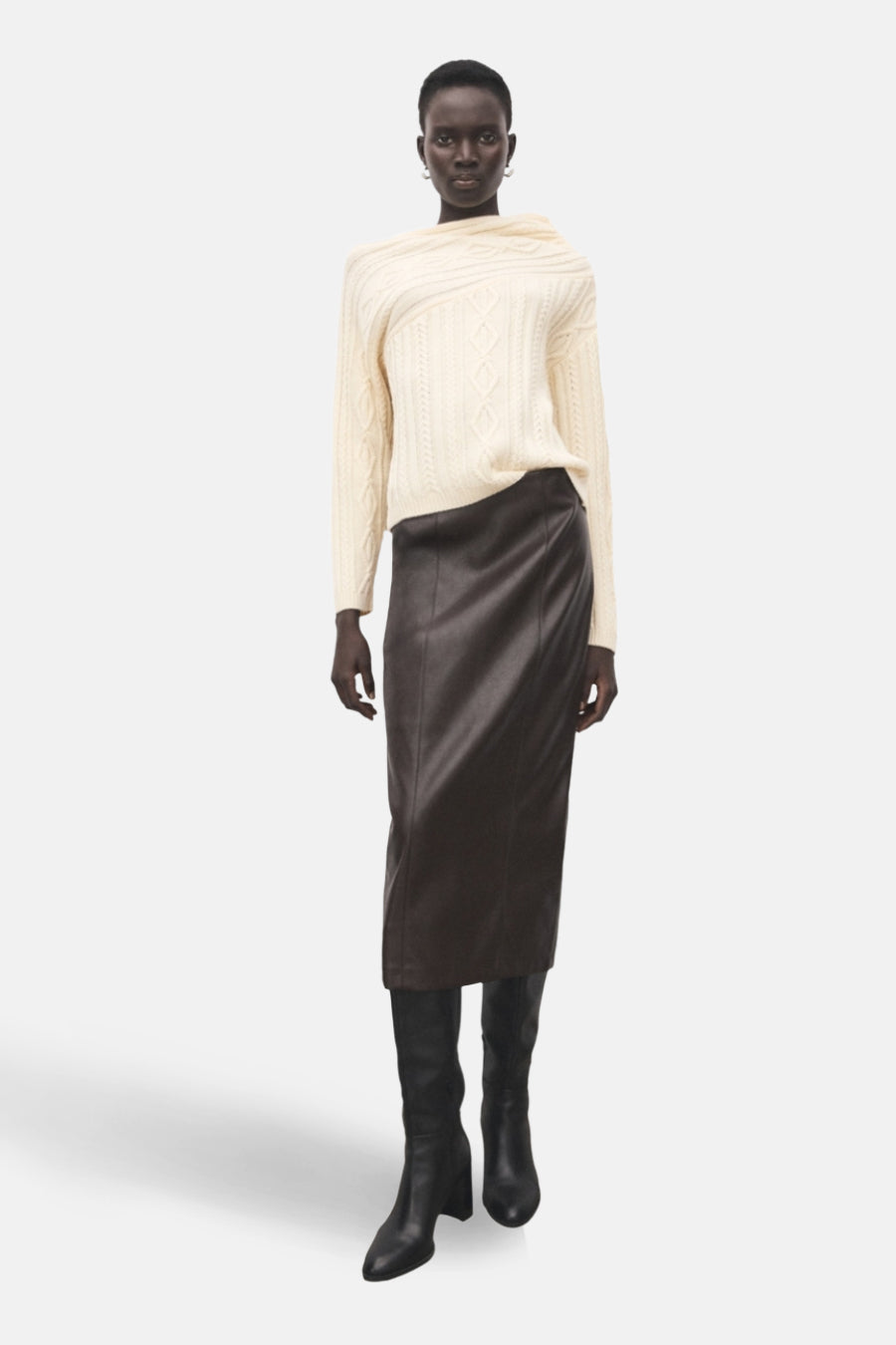 Pull à col rond écru de MANGO, avec un motif torsadé et porté avec une jupe en cuir marron et des bottes noires.