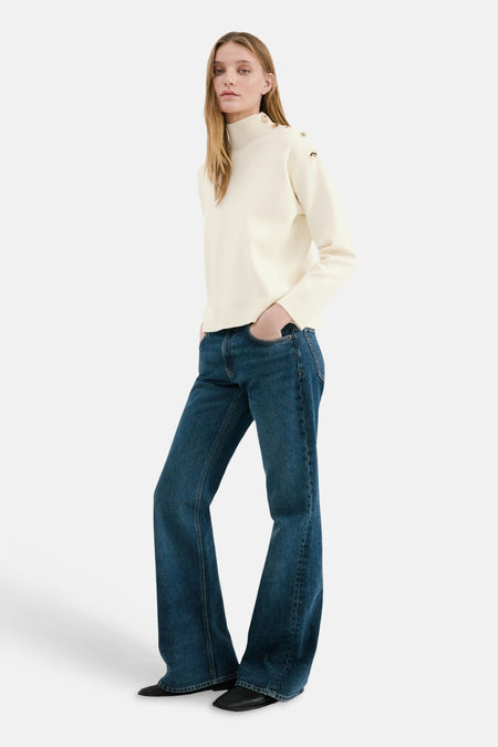 Pull à col rond écru de MANGO, avec des boutons décoratifs aux épaules, porté avec un jean évasé et des chaussures noires.
