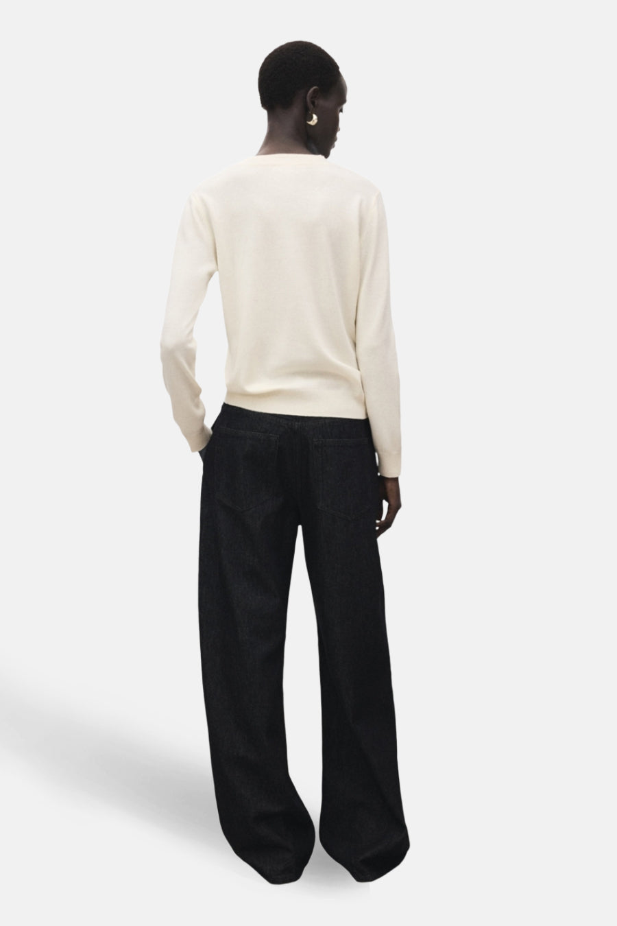 Pull à col rond écru de MANGO, vue de dos, porté avec un pantalon large noir et des boucles d'oreilles dorées.