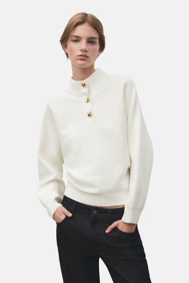 Pull à col roulé écru de MANGO, avec trois boutons dorés, porté avec un jean noir.
