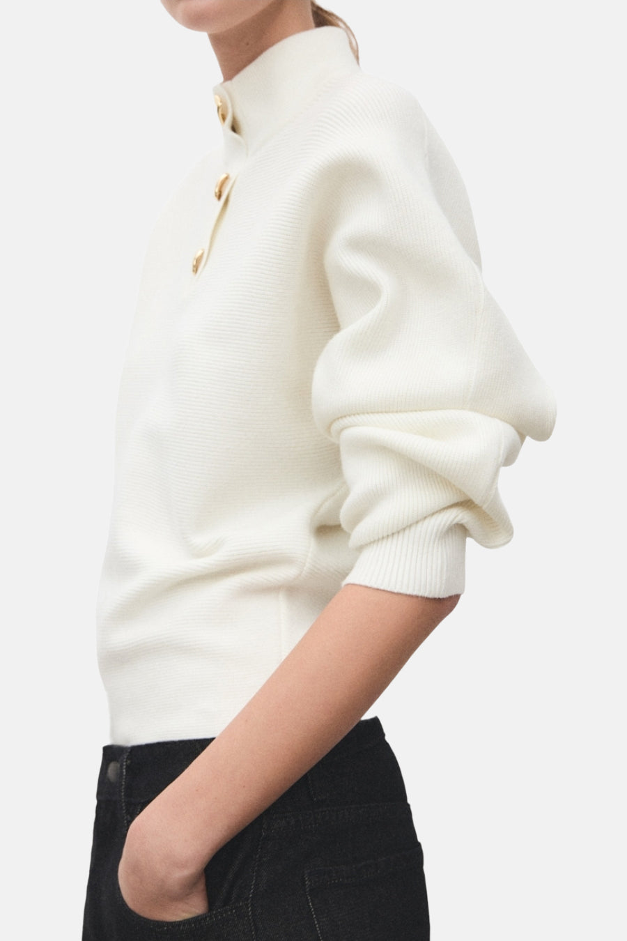 Pull à col roulé écru de MANGO, avec boutons décoratifs dorés, porté avec un pantalon noir, en vue de côté.
