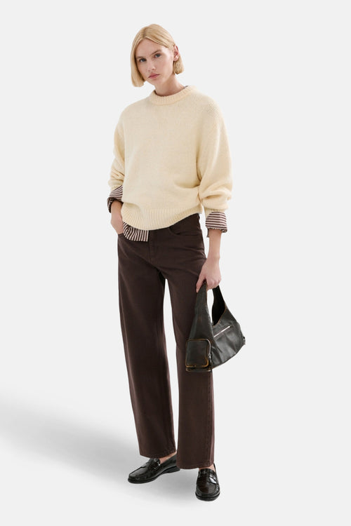 Jean coloré brun de MANGO, porté avec un pull crème, une chemise rayée et des mocassins.
