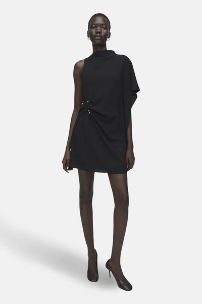 Robe noire asymétrique de MANGO, avec drapé à la taille et finitions minimalistes, portée avec des escarpins noirs classiques.
