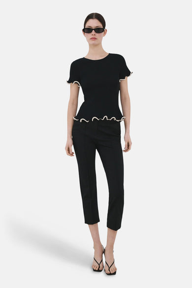 Pull col rond noir de MANGO, avec ourlet ondulé, porté avec un pantalon noir et des sandales à brides.