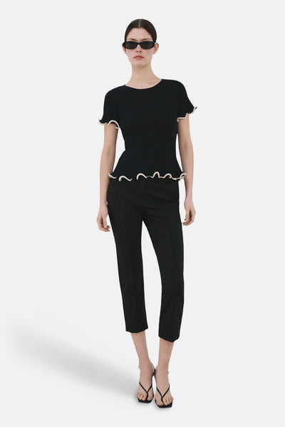 Pull col rond noir de MANGO, avec ourlet ondulé, porté avec un pantalon noir et des sandales à brides.