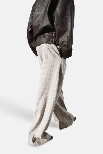 Beige broek van MANGO, met discrete ruitjes, wijde pasvorm en vallend over donkere schoenen, gedragen met een bruin leren jasje.