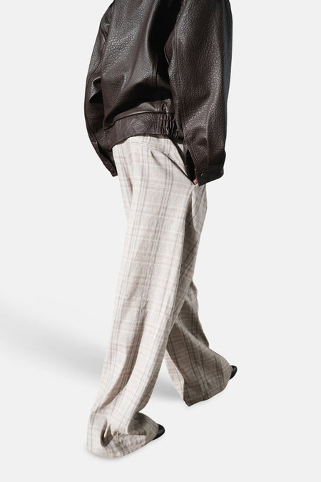 Pantalon beige de MANGO, à carreaux discrets, coupe ample et tombant sur des chaussures sombres, porté avec une veste en cuir marron.
