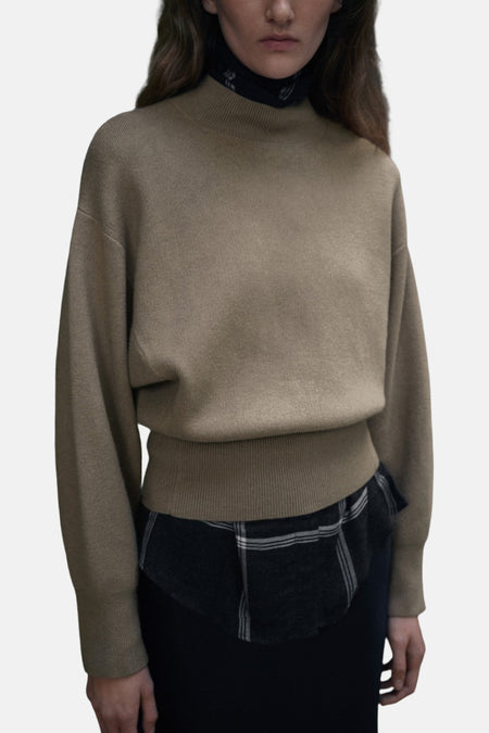Pull à col rond kaki de MANGO, porté sur une chemise à carreaux et une jupe foncée.
