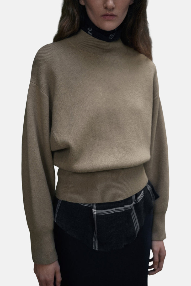 Pull à col rond kaki de MANGO, porté sur une chemise à carreaux et une jupe foncée.
