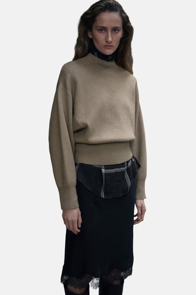 Pull à col rond kaki de MANGO, porté avec une chemise à carreaux et une jupe noire à dentelle.
