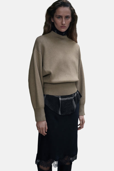 Pull à col rond kaki de MANGO, porté avec une chemise à carreaux et une jupe noire à dentelle.
