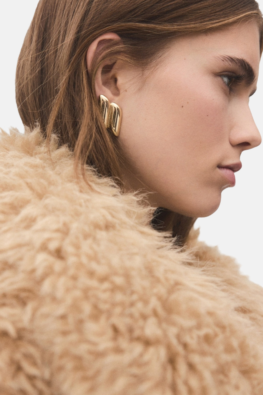 Boucles d'oreilles dorées de MANGO, design ondulé, portées avec un manteau texturé couleur crème.