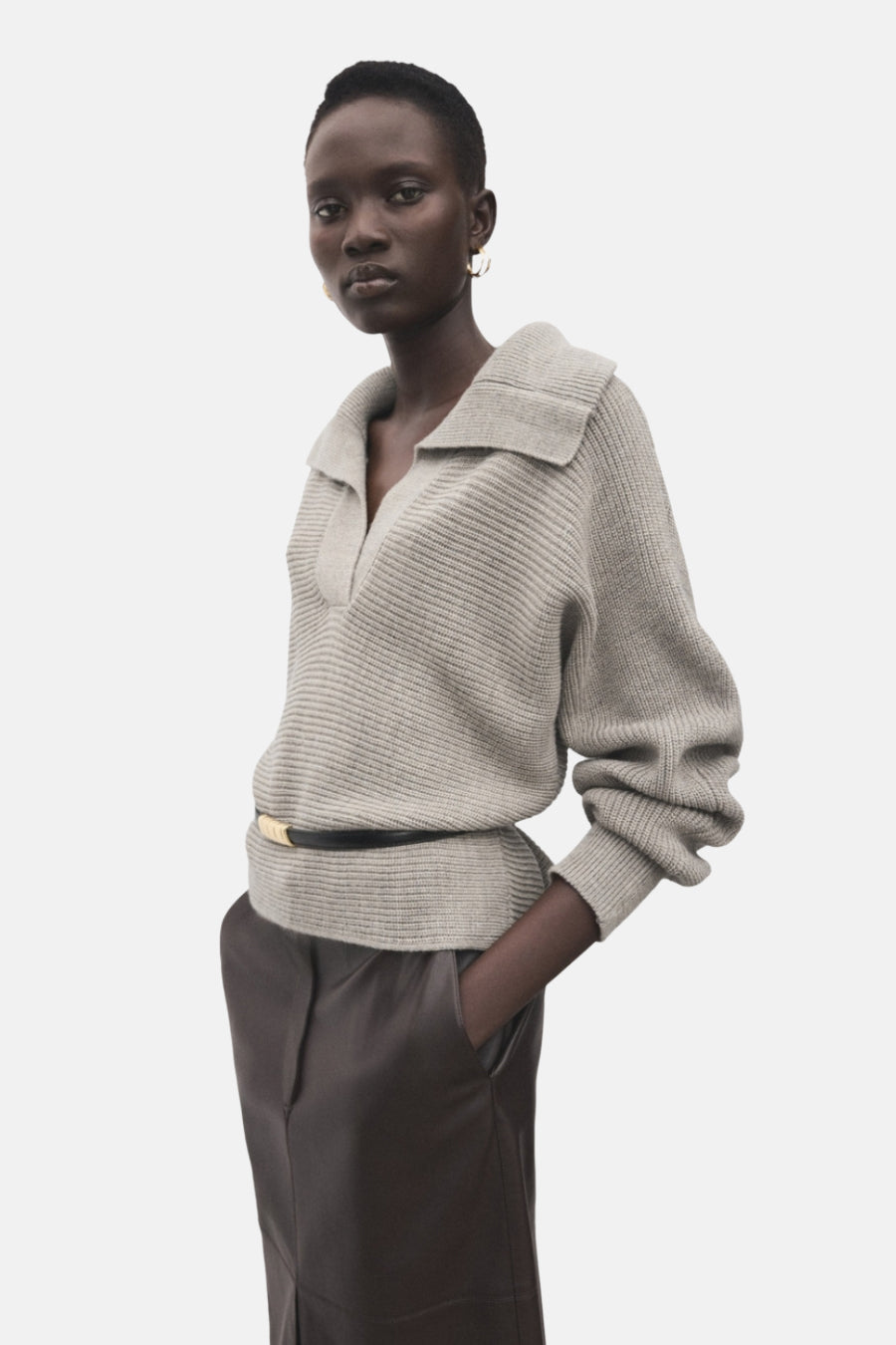 Pull à col en V beige de MANGO, porté avec une ceinture noire à boucle dorée et un pantalon brun.