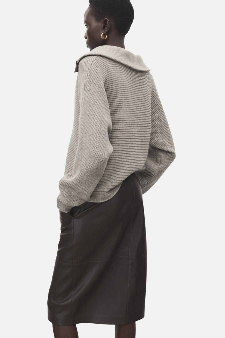 Pull à col en V beige de MANGO, avec une jupe en cuir marron et des boucles d'oreilles dorées.