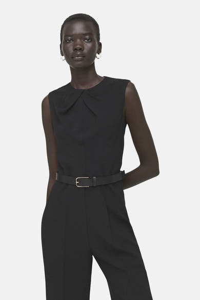 Zwarte jumpsuit van MANGO, met riem en ronde hals, gedragen door een model met oorbellen.