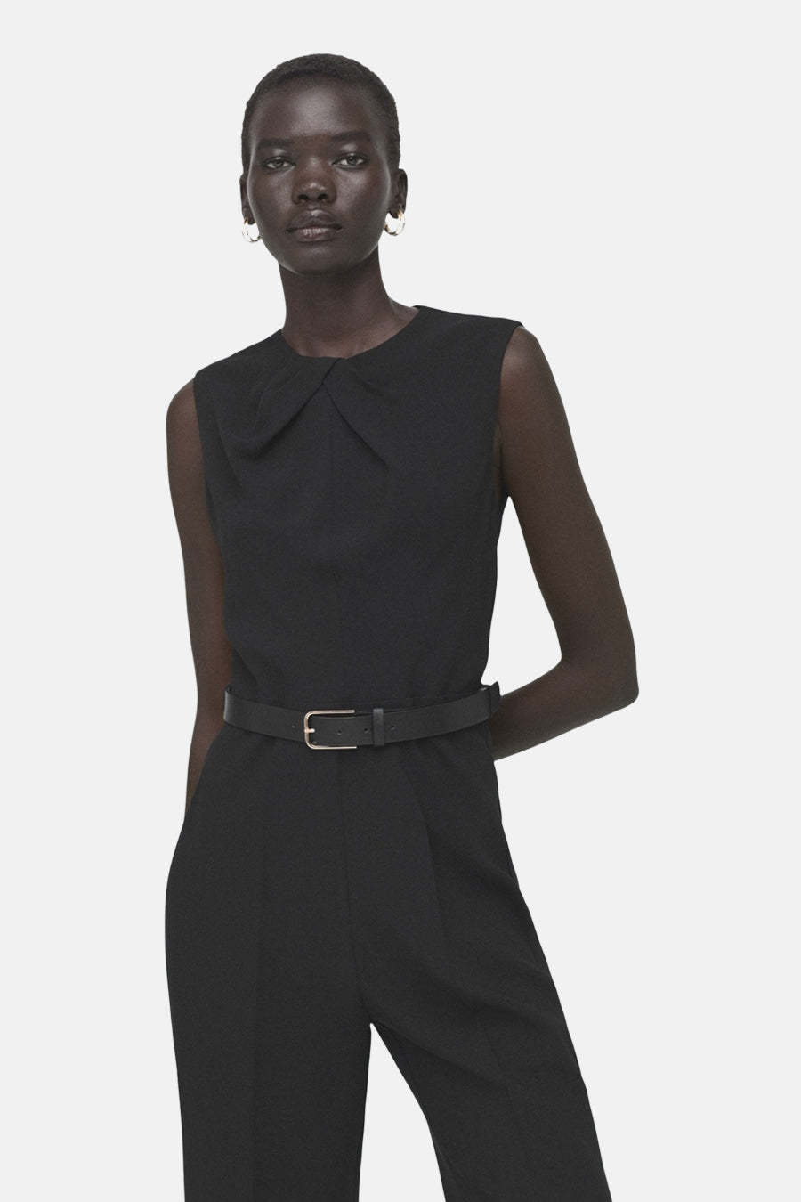 Zwarte jumpsuit van MANGO, met riem en ronde hals, gedragen door een model met oorbellen.