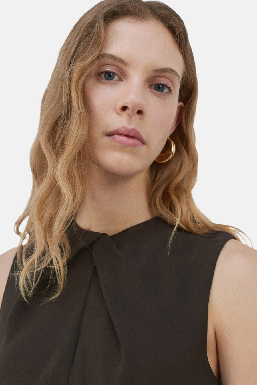 Combinaison brune de MANGO avec un col croisé, portée avec des boucles d'oreilles dorées et des cheveux ondulés blonds.
