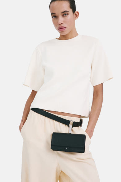Pull col rond écru de MANGO, porté avec un pantalon couleur crème et une ceinture avec un petit sac attaché.

