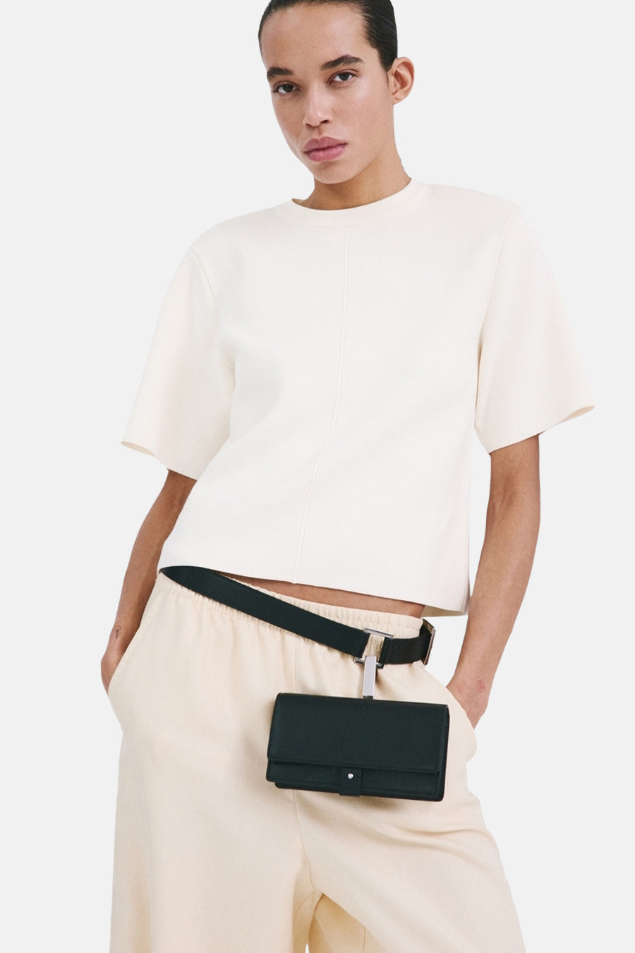 Pull col rond écru de MANGO, porté avec un pantalon couleur crème et une ceinture avec un petit sac attaché.
