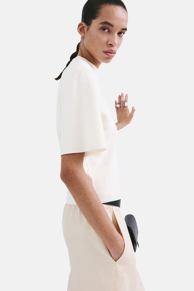 Pull écru à col rond de MANGO, manches courtes amples et détail de ceinture noire, porté avec un pantalon beige.
