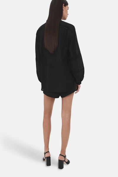 Chemisier noir à manches longues de MANGO, avec des détails ajourés et des finitions soignées, porté avec des shorts et des chaussures à talons noirs.