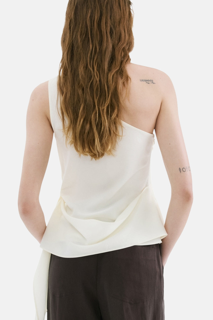Blouse blanche asymétrique de MANGO, drapée, portée de dos avec un pantalon marron.