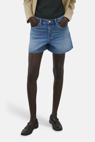 Bermuda en denim bleu moyen de MANGO, porté avec des mocassins en cuir marron et une veste beige.