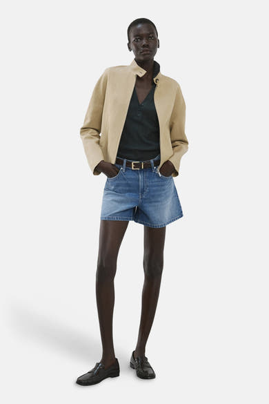 Middelblauwe denim bermuda van MANGO, gedragen met een beige jasje, een donkergroen poloshirt en zwarte loafers.