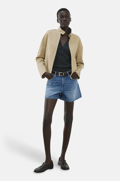 Bermuda en denim bleu moyen de MANGO, porté avec une veste beige, un polo vert foncé et des mocassins noirs.
