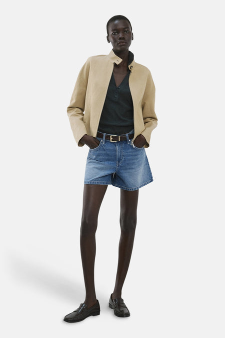 Bermuda en denim bleu moyen de MANGO, porté avec une veste beige, un polo vert foncé et des mocassins noirs.
