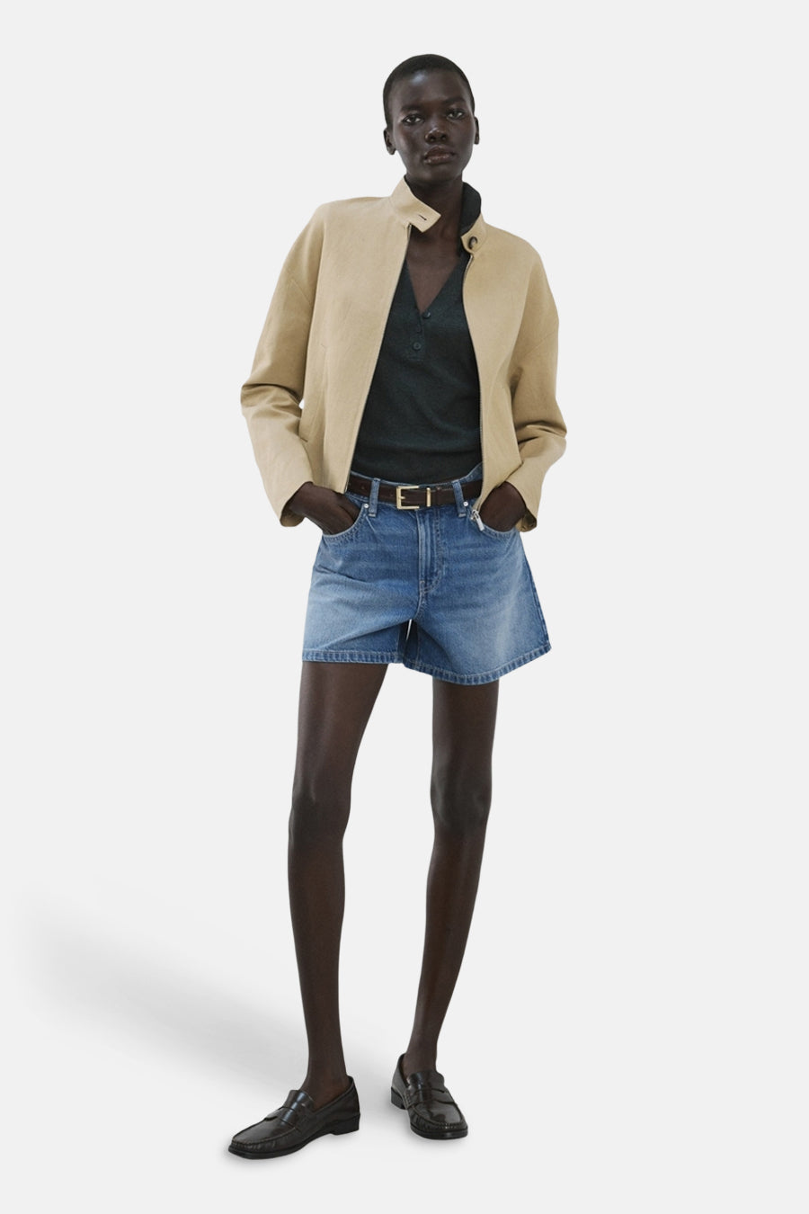 Middelblauwe denim bermuda van MANGO, gedragen met een beige jasje, een donkergroen poloshirt en zwarte loafers.