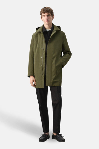 Lange kaki jas van MANGO, met capuchon, gedragen over een zwart overhemd en een zwarte broek, model ten voeten uit.