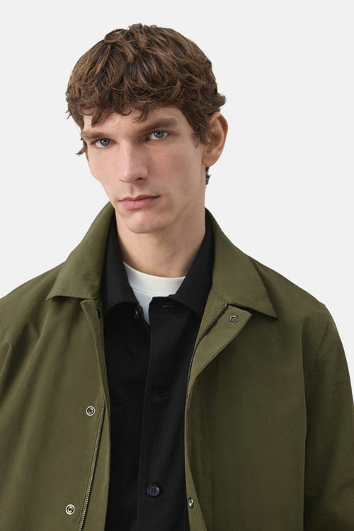 Veste longue kaki de MANGO, portée avec une chemise noire et un t-shirt blanc, sur fond clair.