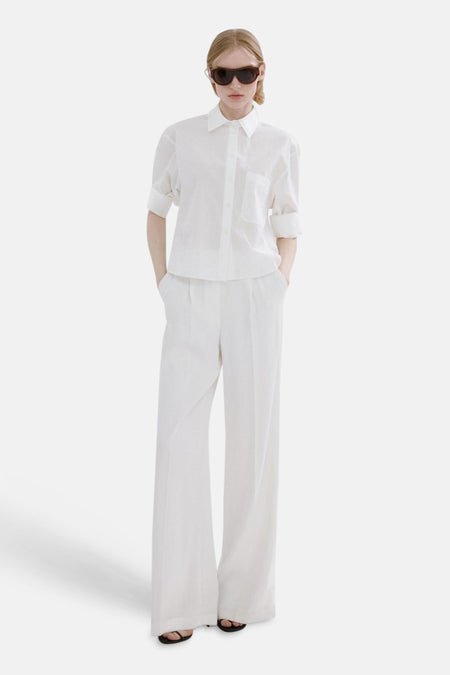 Chemise à manches longues blanche de MANGO, avec poche poitrine et manches retroussées, portée avec pantalon large et lunettes de soleil.
