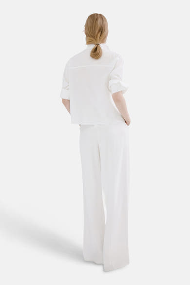 Chemisier à manches longues blanc de MANGO, porté avec un pantalon blanc, vue de dos, cheveux blonds attachés.