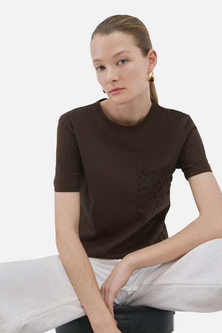 T-shirt brun de MANGO avec détail brodé ton sur ton, porté avec un pantalon blanc et des boucles d'oreilles dorées.