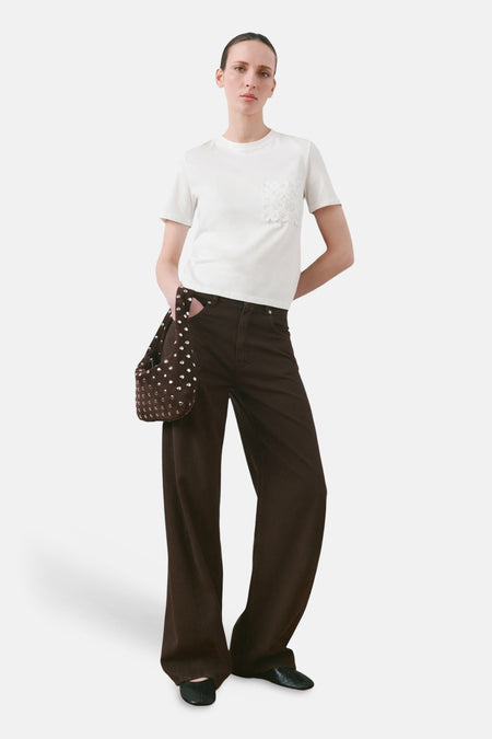 T-shirt blanc à manches courtes de MANGO, avec poche en dentelle sur la poitrine. Porté avec un pantalon marron ample et un sac à main.