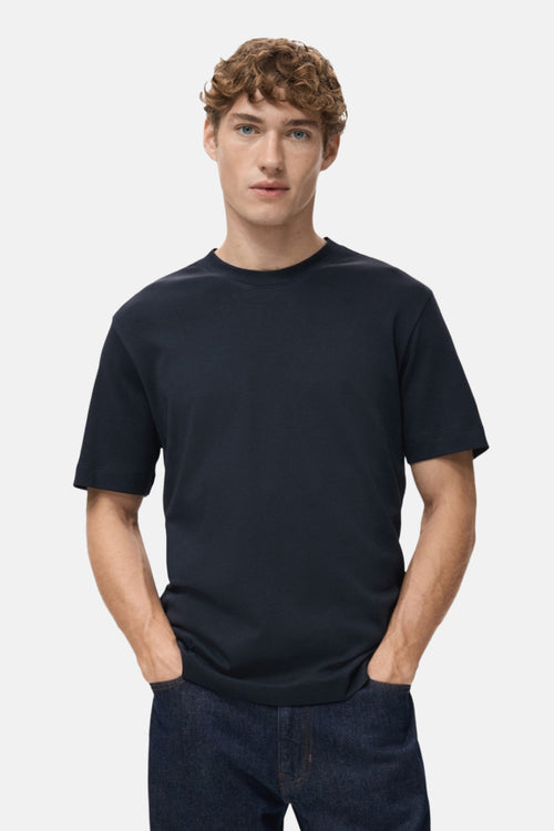 T-shirt à manches courtes bleu de MANGO, porté avec un jean bleu foncé et les mains dans les poches.
