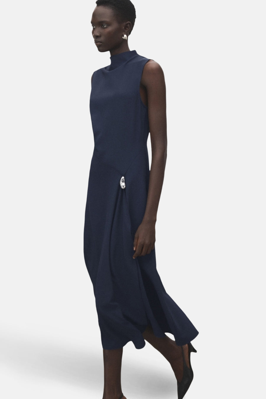 Blauwe jurk van MANGO, met een asymmetrische snit, versierd met een decoratieve knoop, gedragen met zwarte pumps.