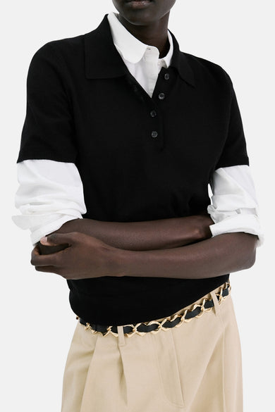 Polo noir à manches courtes de MANGO, porté sur une chemise blanche avec un pantalon taille haute beige et une ceinture chaîne dorée.
