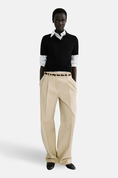 Polo noir à manches courtes de MANGO, porté sur une chemise blanche, avec un pantalon beige et une ceinture dorée.

