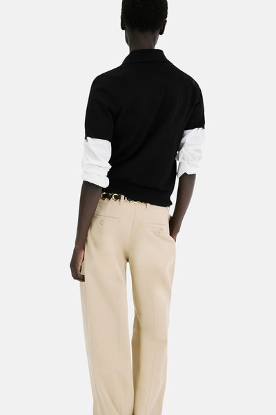 Polo à manches courtes noir de MANGO, porté avec une chemise blanche à manches longues et un pantalon beige avec ceinture dorée.