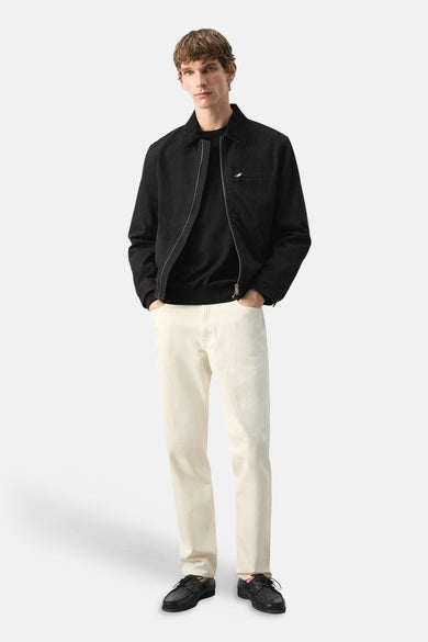 Veste courte noire par MANGO, avec fermeture éclair et poches poitrine, portée avec un pull noir et un jean écru.
