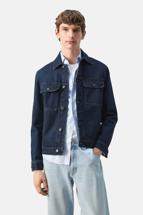 Veste en jean en denim bleu foncé de MANGO, portée sur une chemise rayée, avec un pantalon en jean clair.
